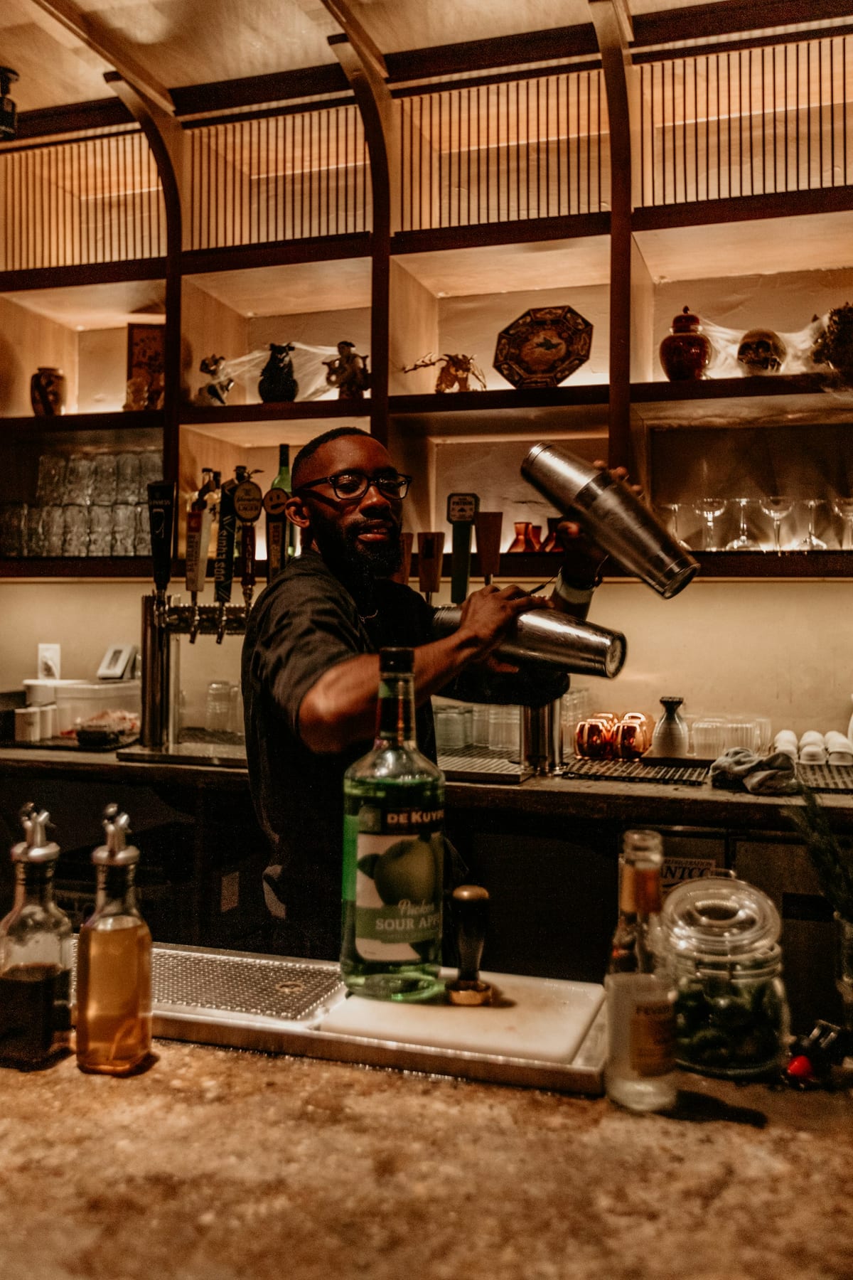 A bartender shaking a cocktail at a back bar lined with bottles
